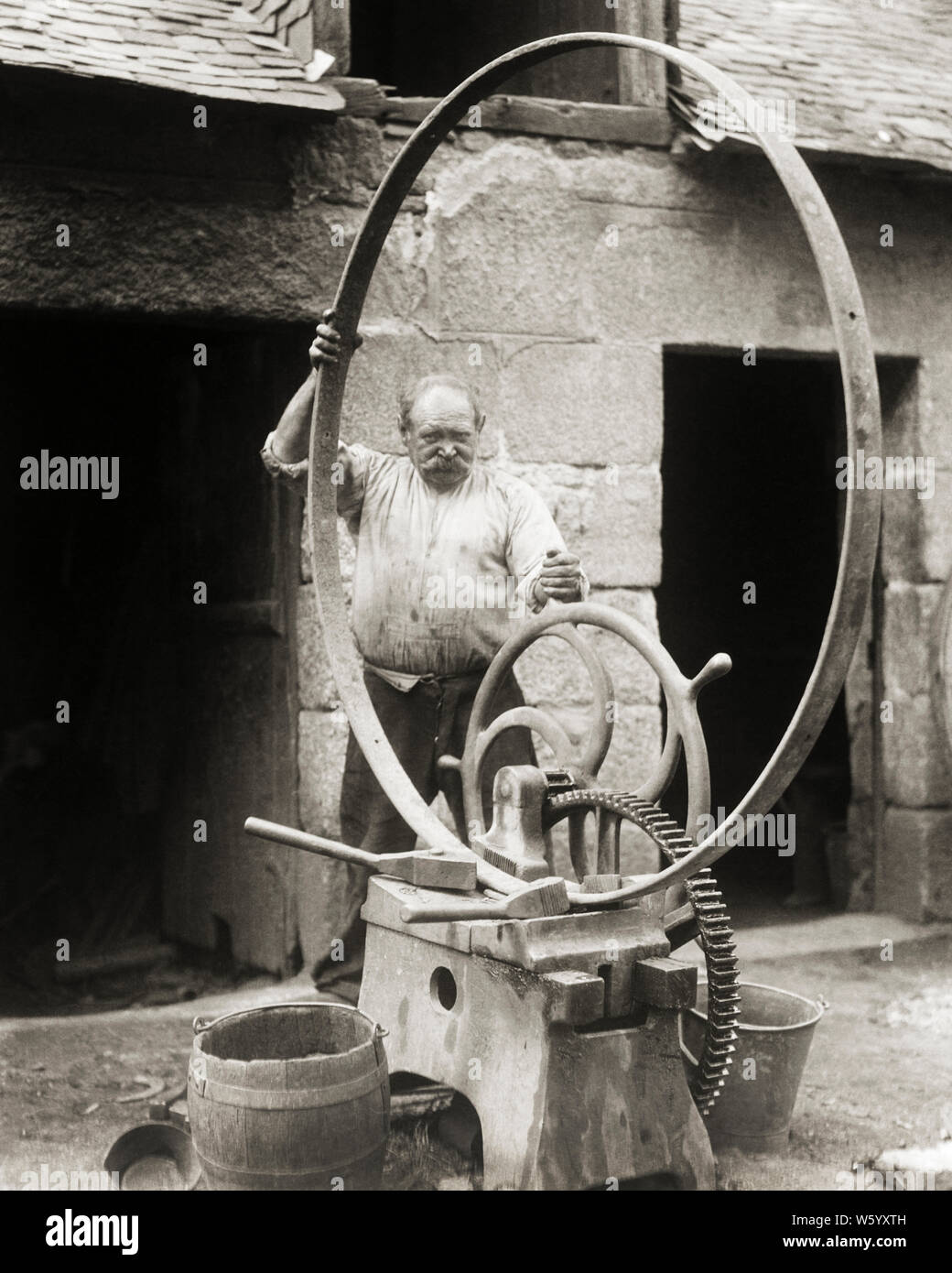 1920s MAN MILLWRIGHT OR BLACKSMITH BENDING LARGE ROUND IRON HOOP REINFORCING BAND FOR A WINE STORAGE BARREL BRITTANY FRANCE  - r4423 HAR001 HARS PROFESSION CRAFT BARREL SHAPE EUROPE MIDDLE-AGED B&W MIDDLE-AGED MAN BENDING SKILL OCCUPATION SKILLS GEARS EUROPEAN MACHINERY STRENGTH CAREERS EXTERIOR KNOWLEDGE POWERFUL BLACKSMITH LABOR A AUTHORITY EMPLOYMENT OCCUPATIONS CIRCULAR STYLISH EMPLOYEE OR BRITTANY CRAFTSMAN CREATIVITY REINFORCING BLACK AND WHITE CAUCASIAN ETHNICITY HAR001 LABORING OLD FASHIONED Stock Photo