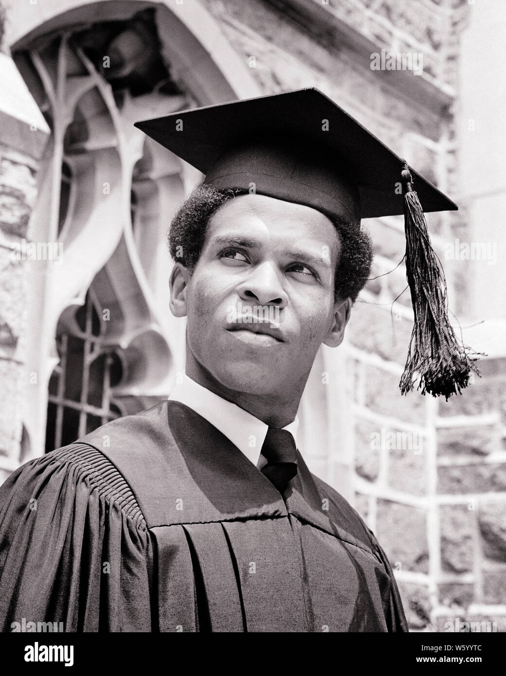 1970s AFRICAN-AMERICAN HIGH SCHOOL GRADUATE WEARING CAP AND GOWN - s18239 HAR001 HARS KNOWLEDGE LEADERSHIP LOW ANGLE BLACK ETHNICITY PRIDE OPPORTUNITY HIGH SCHOOL HIGH SCHOOLS HIGHER EDUCATION CONCEPTUAL COLLEGES MORTARBOARD GROWTH YOUNG ADULT MAN BLACK AND WHITE CAP AND GOWN HAR001 OLD FASHIONED AFRICAN AMERICANS Stock Photo