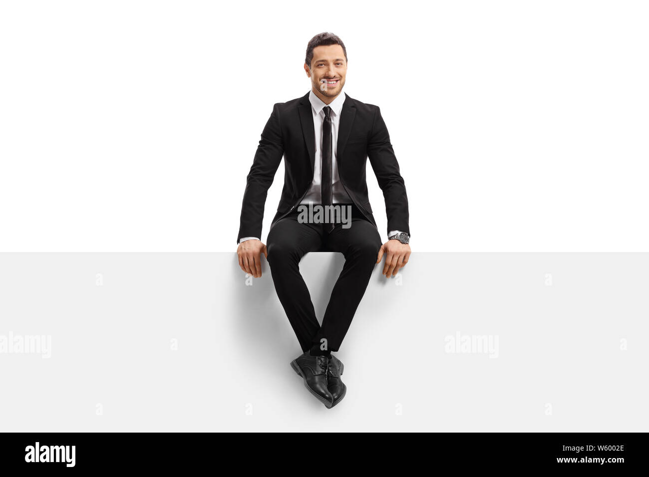 Full length portrait of a young man in a suit sitting on a panel and smiling isolated on white background Stock Photo