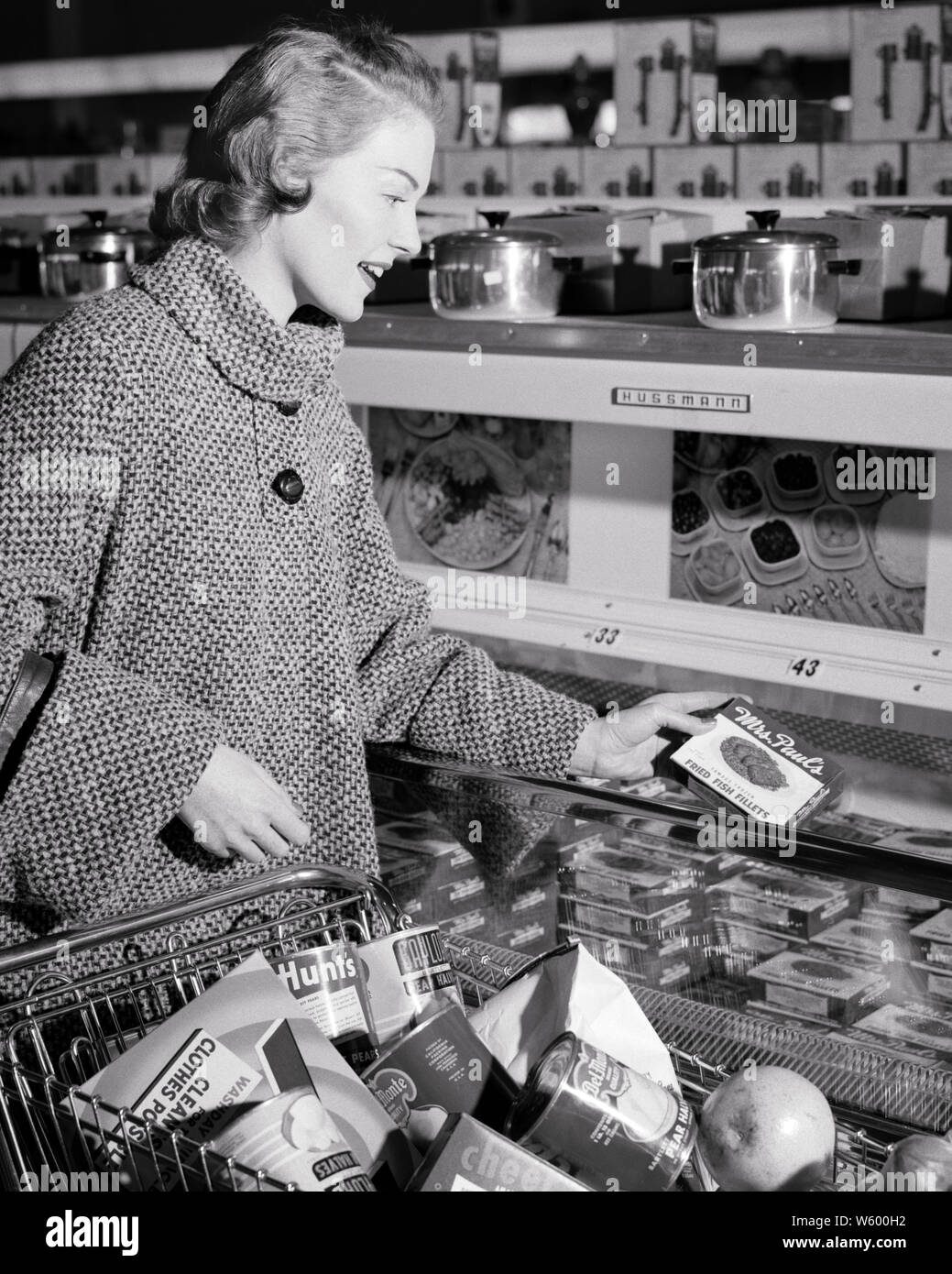 WOMAN SEARING COAT SHOPPING IN FROZEN FOOD SECTION OF SUPERMARKET - s5920 HAR001 HARS HOMEMAKER SHOPPERS HAPPINESS HOMEMAKERS CHEERFUL HIGH ANGLE KART SHOPPING CART CARTS HOUSEWIVES SMILES JOYFUL STYLISH MID-ADULT MID-ADULT WOMAN BLACK AND WHITE CAUCASIAN ETHNICITY HAR001 OLD FASHIONED SECTION Stock Photo