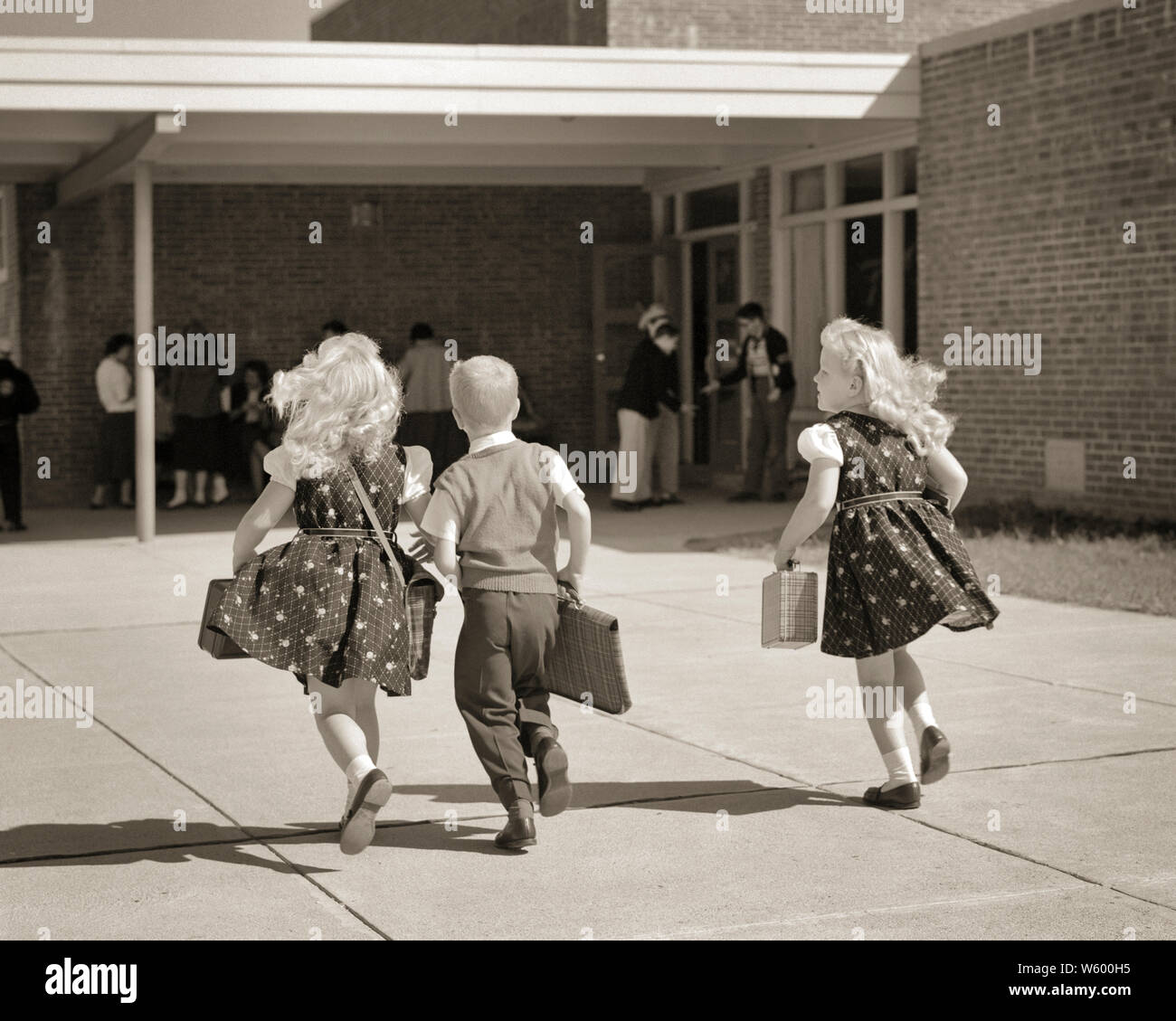 1950s BOY AND TWO TWIN GIRLS RUNNING TO SCHOOL ENTRANCE BACK VIEW CARRYING LUNCH BOXES BOOK BAGS GIRLS WEARING MATCHING DRESSES - s6894 HAR001 HARS FRIEND TEAMWORK TWIN IDENTICAL DOUBLE JOY LIFESTYLE FEMALES BROTHERS COPY SPACE FRIENDSHIP FULL-LENGTH MATCH MALES SIBLINGS SISTERS B&W LATE DRESSES MATCHING SAME SCHOOLS GRADE AND EXCITEMENT KNOWLEDGE REAR VIEW PRIMARY SIBLING CONNECTION LUNCH BOXES FRIENDLY STYLISH K-12 LOOK-ALIKE BACK VIEW COOPERATION DUPLICATE GRADE SCHOOL GROWTH JUVENILES LOOK ALIKE TARDY TOGETHERNESS BLACK AND WHITE CAUCASIAN ETHNICITY CLONE HAR001 HURRY OLD FASHIONED Stock Photo