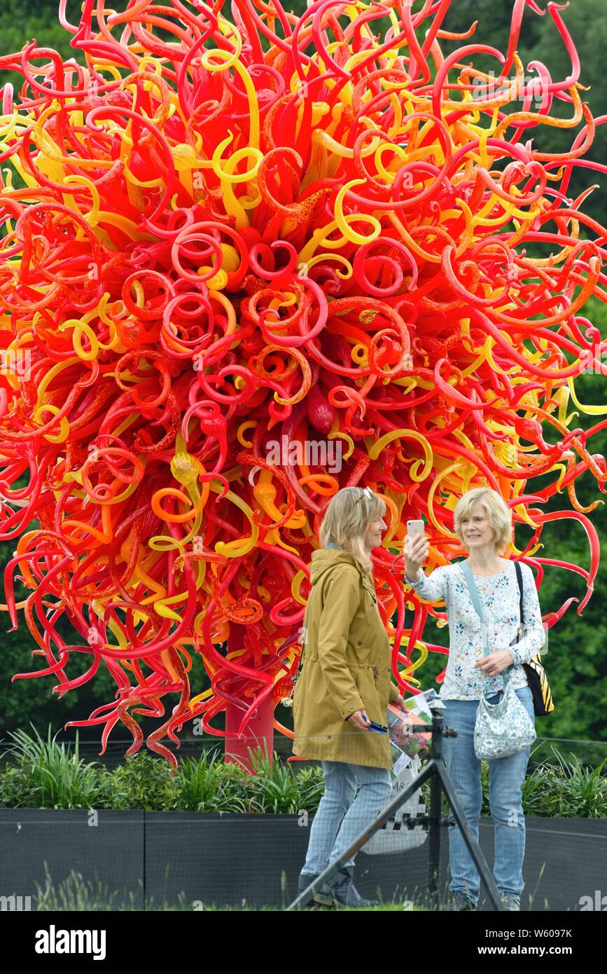 Two women taking selfies against  a spectacular glass sculpture by Dale Chihuly on display at Kew Botanical Gardens West London England UK Stock Photo