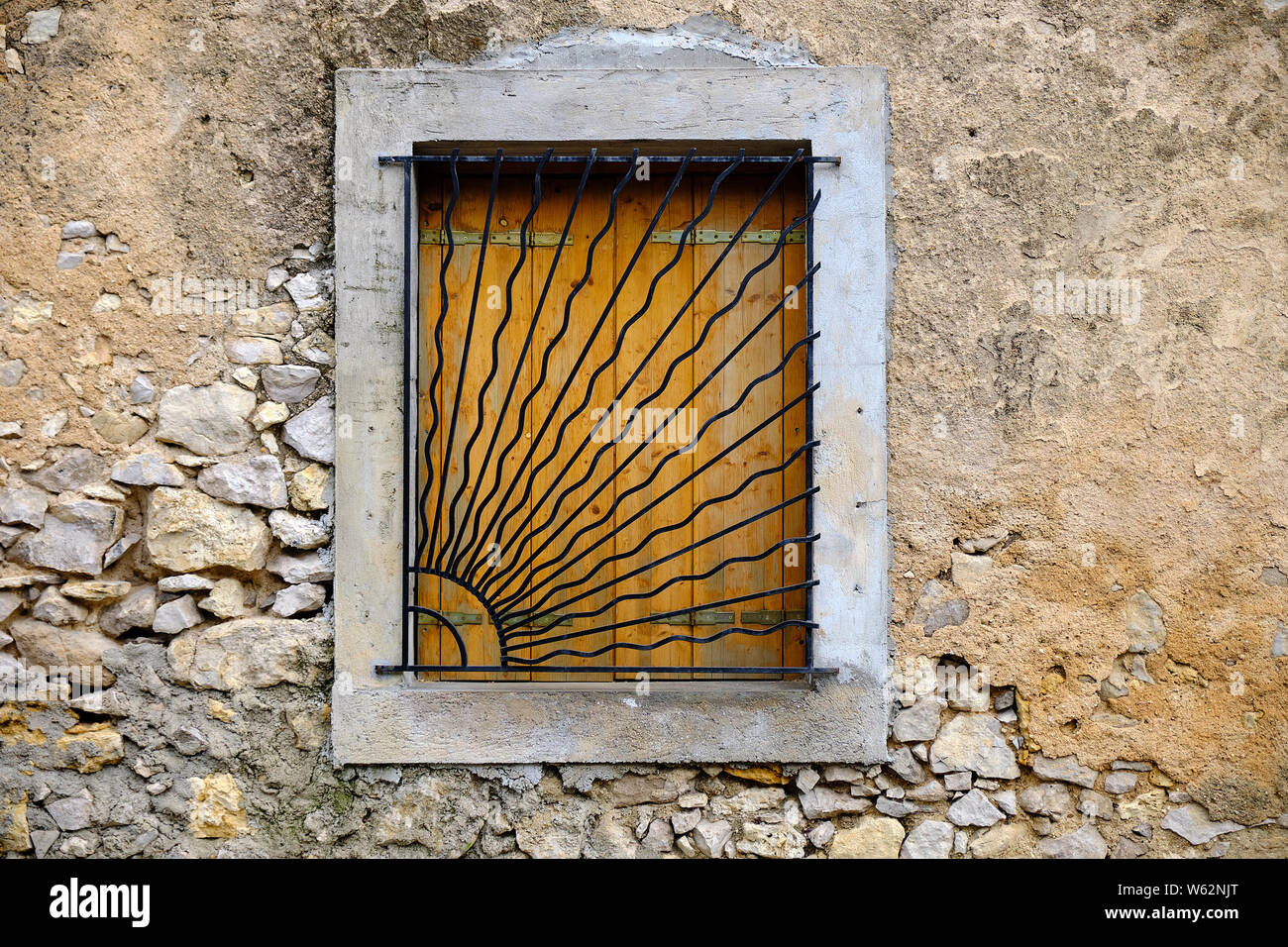 Old house window with metal bars in shape of morning sun closed shutter in textured rocks wall Stock Photo