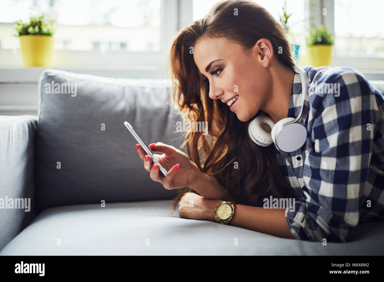 Side view of a young attractive woman smiling and looking at phone while lying on sofa Stock Photo