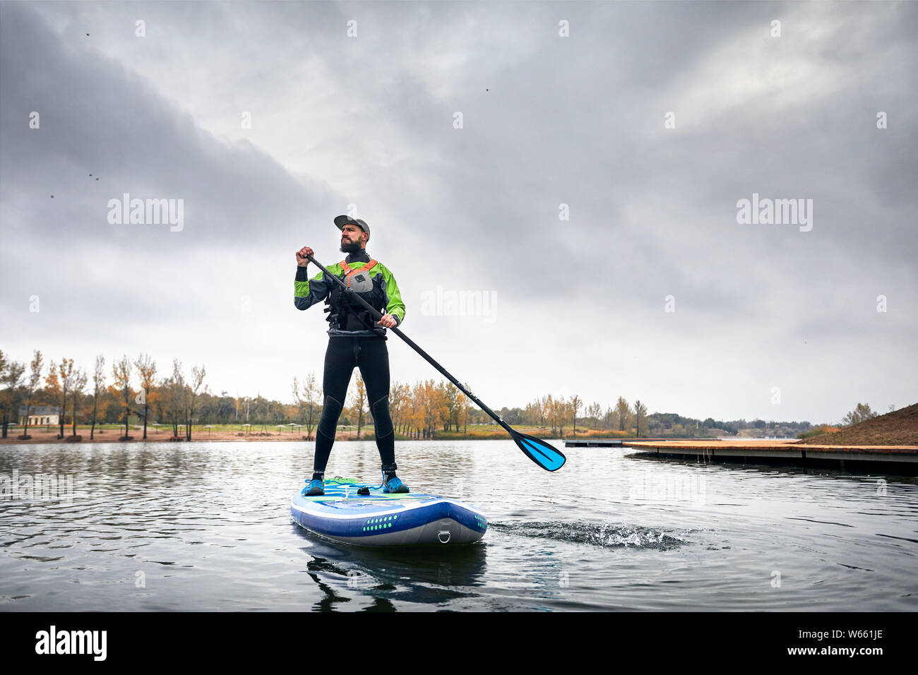 Athlete in wetsuit on paddleboard exploring the lake at cold weather against overcast sky Stock Photo