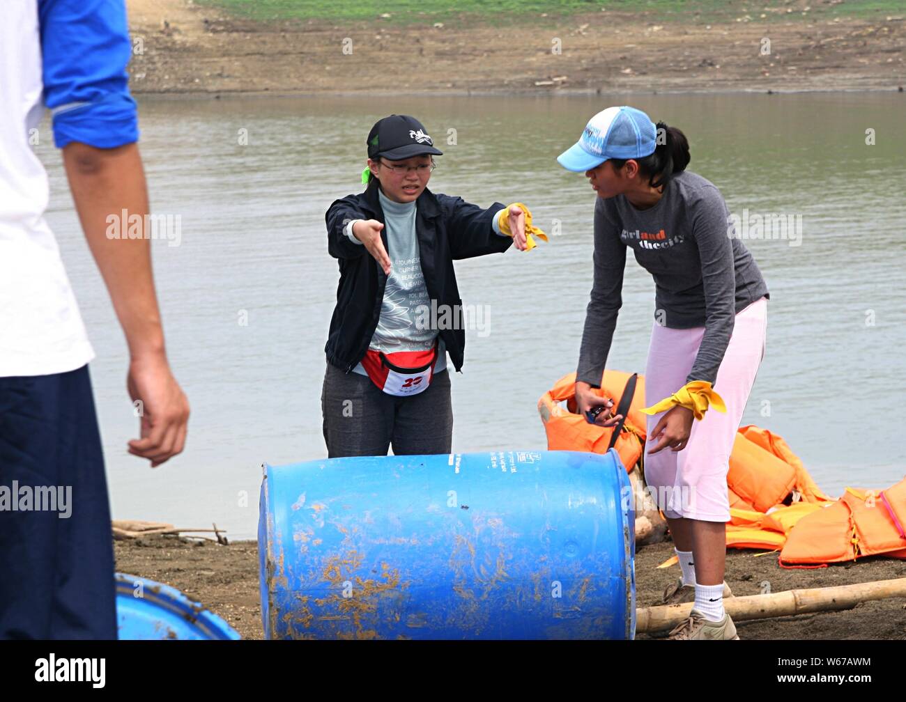 A young woman give instruction to her friend about making a boat using plastic barrels, bamboo sticks and ropes beside a water dam. Stock Photo