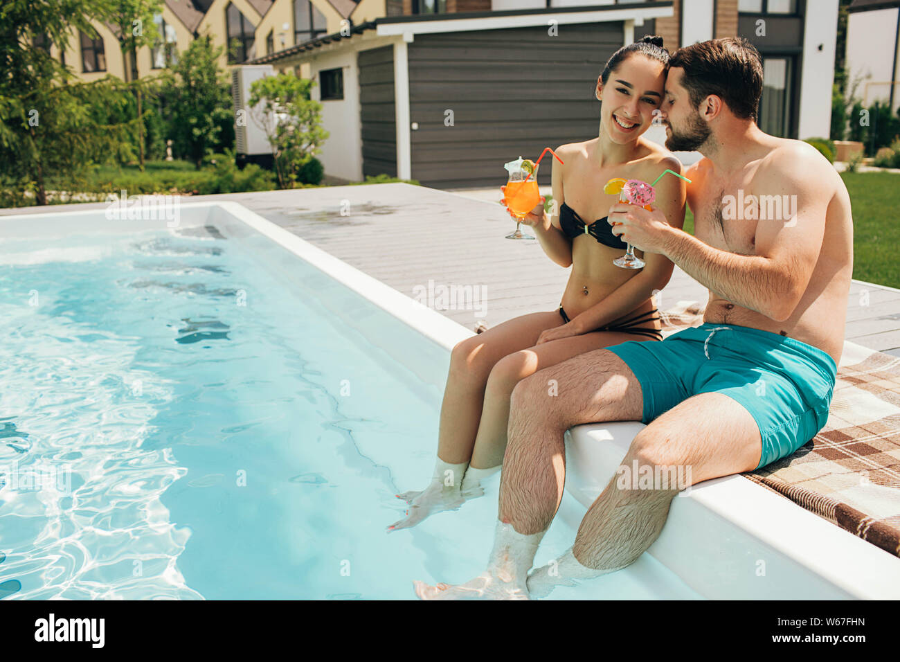 Happy couple sitting by the poolside in the backyard of their house. Man and woman sitting near water pool and funny splashing water their legs Stock Photo