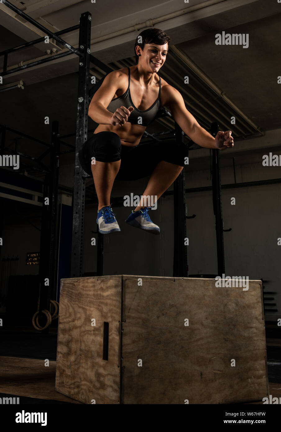 An athletic short haired woman with strong abs is doing box jumps in a gym. The female athlete is smiling and jumping on the box. Black and white. Stock Photo