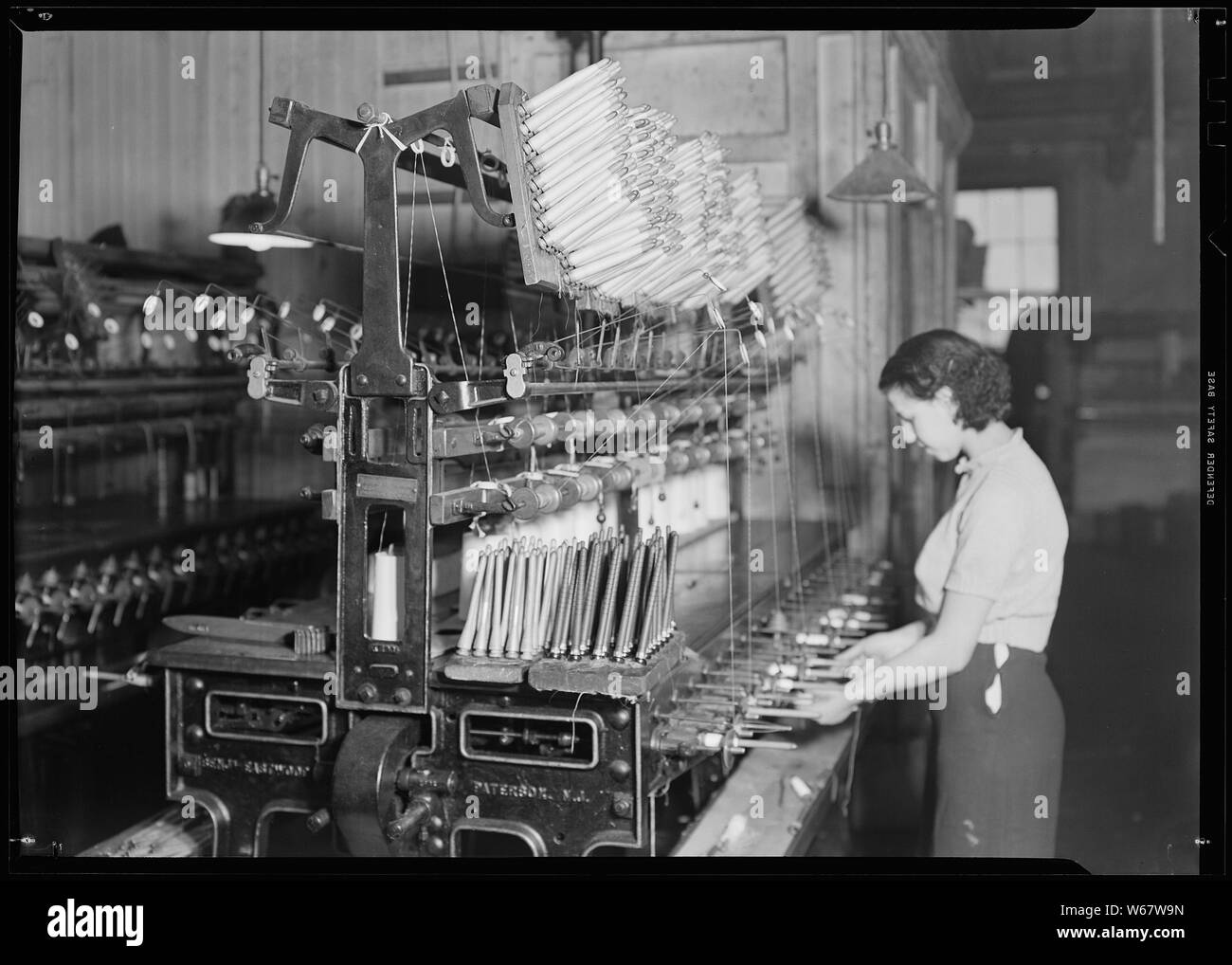 Paterson, New Jersey - Textiles. Quilling. The thread is wound from the cones, seen in an upright position on the table of the machine, on to the quills soon at the position of the operator's hands. Here the rack of partially filled quills hanging on the top of the machine and the racks of empty quills on the table of the machine. Stock Photo