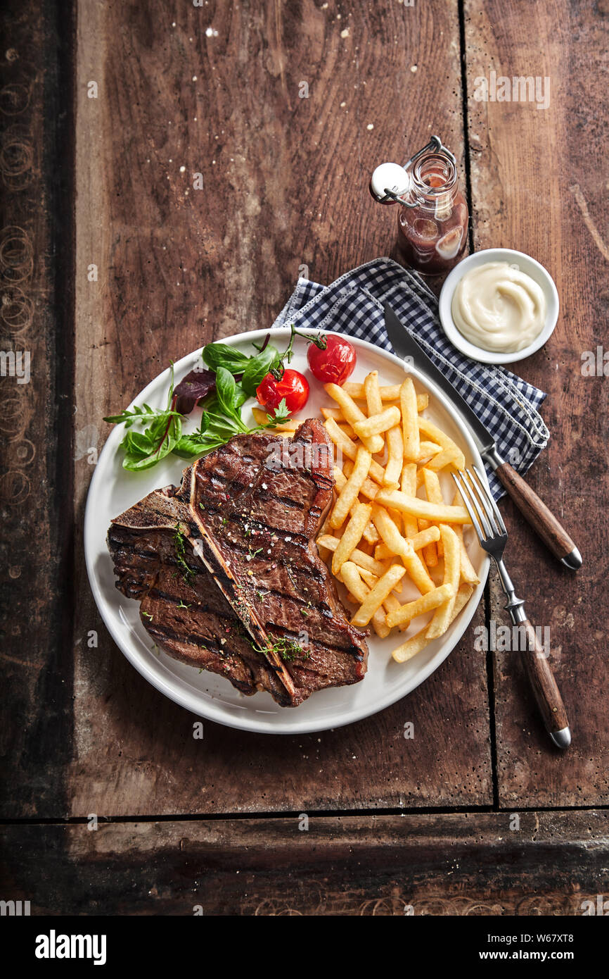 Grilled lean T-bone steak with golden potato chips and salad served with a dip or dressing on a rustic wooden table in a top down view with copy space Stock Photo
