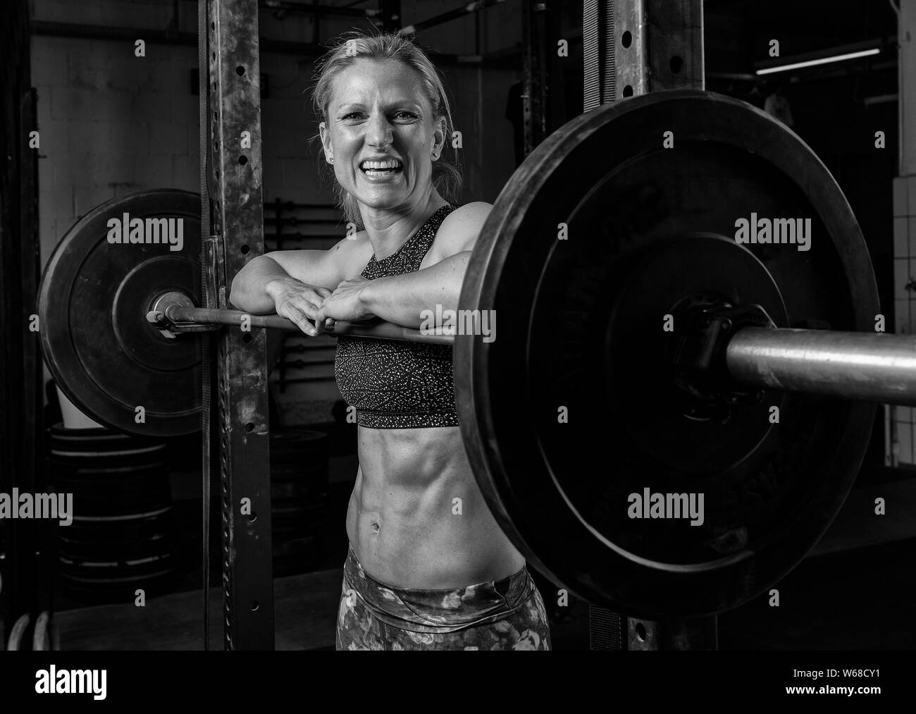 Portrait of a beautiful female athlete with strong abs. The athletic and smiling blonde woman is leaning against the barbells and laughing. Stock Photo