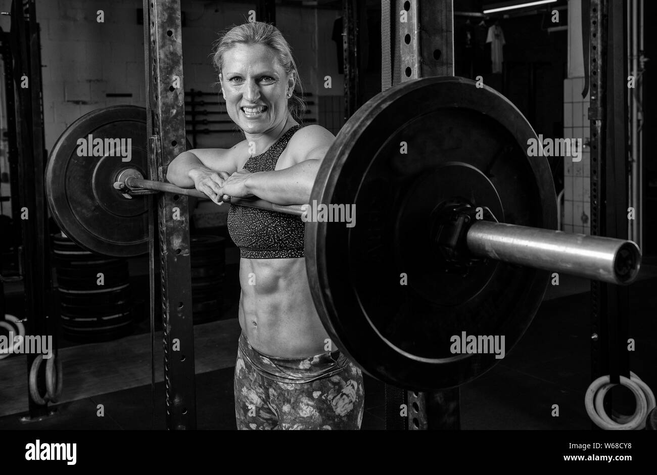 Black and white portrait of an attractive female athlete with strong abs. The athletic and smiling blonde woman is leaning against the barbells. Stock Photo