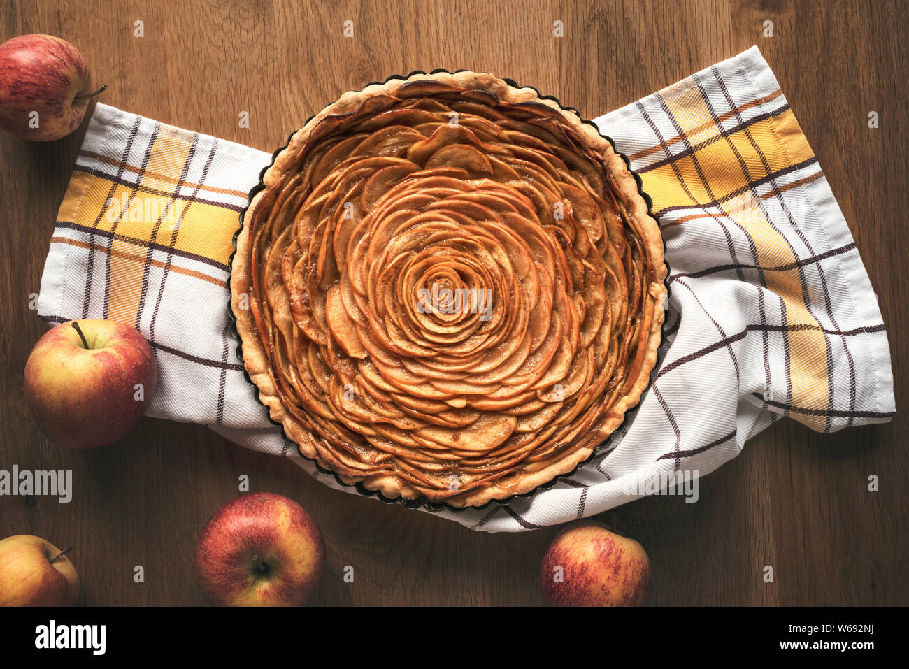 Apple pie decorated in shape of a rose flower, in a tray, on kitchen towel, surrounded by apples fruits, on vintage table. Above view of tatsy apple p Stock Photo