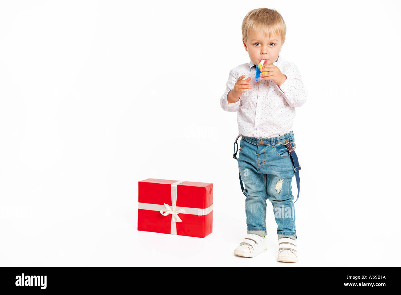 Cute little boy, jeans and shirt, standing near present and blowing in the birthday tune. Isolated on the white background Stock Photo