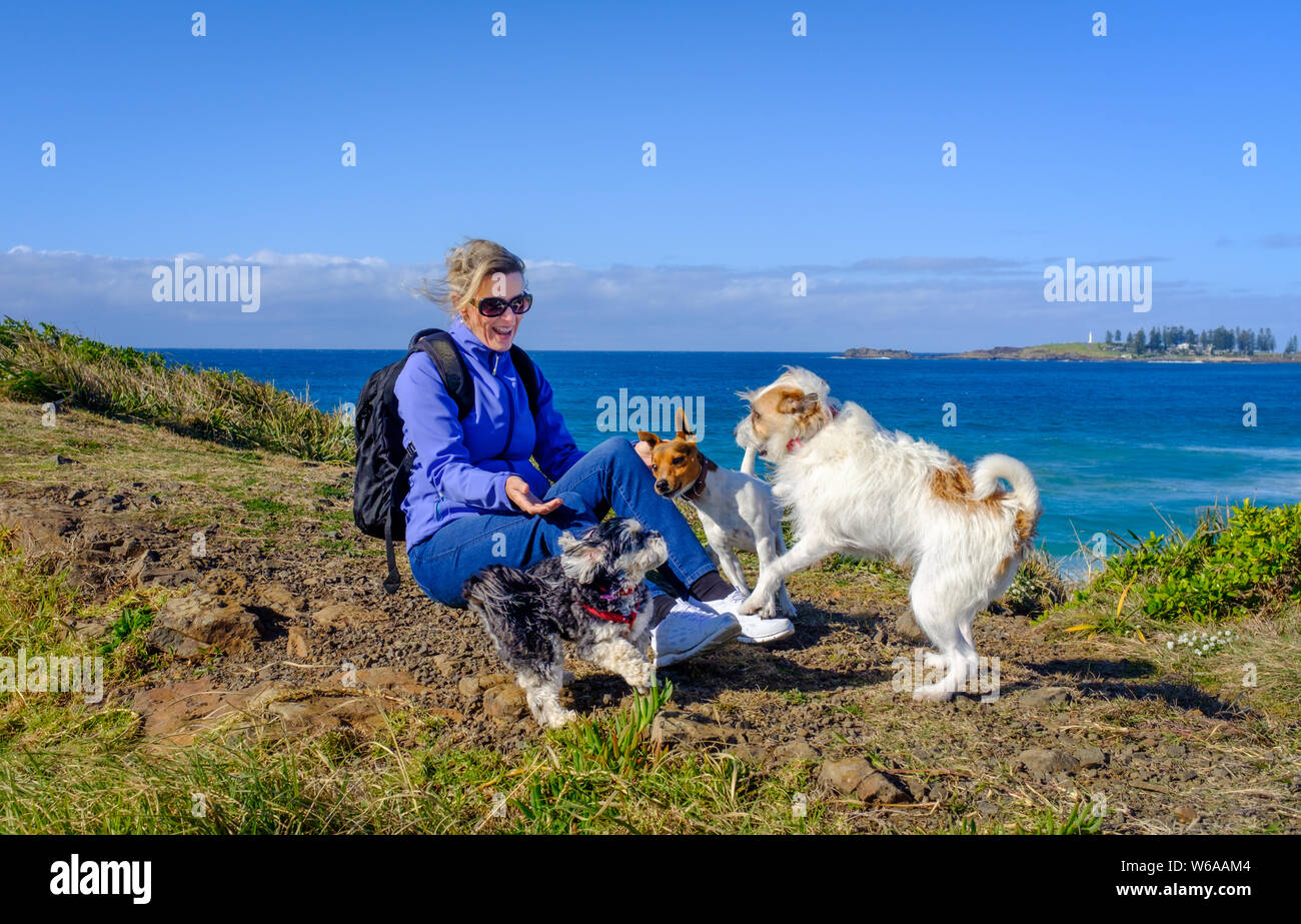 Happy healthy blonde woman 40 plus age in hiking clothes laughing and playing with three cute dogs siting on grass near sea on bright sunny day Stock Photo