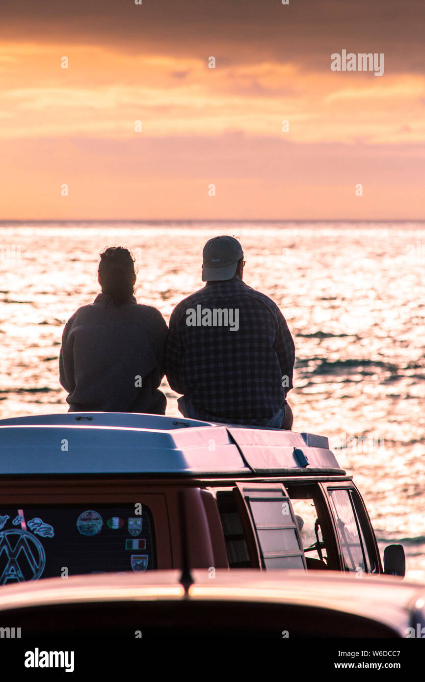A couple sitting on top of their VW Volkswagen Campervan watching a spectacular sunset at Fistral in Newquay in Cornwall. Stock Photo