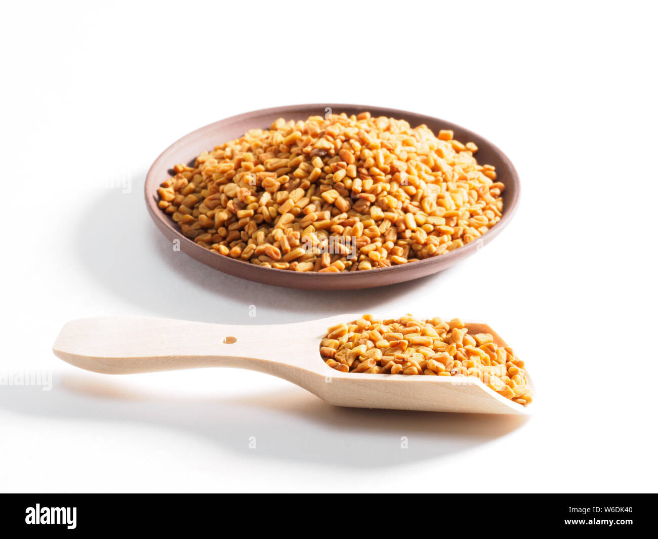 Fenugreek (Trigonetla) on a clay plate with a wooden scoop in the foreground on a white background Stock Photo