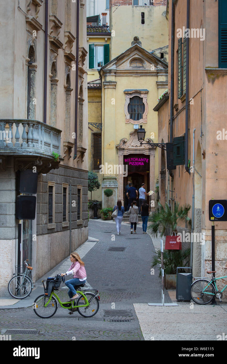 In Verona's old town, a cyclist rides on the Corso Porta Borsari as it crosses the Vicolo St Matteo where the church has become a pizza restaurant. Stock Photo