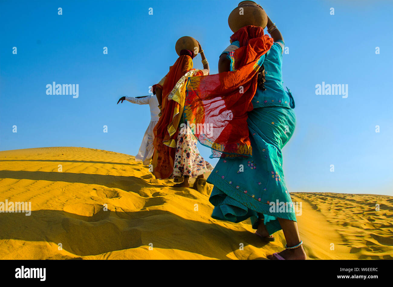 Two women carrying heavy jugs of water on their head and walking on a yellow sand dune in the hot summer desert against blue sky. water crises Stock Photo