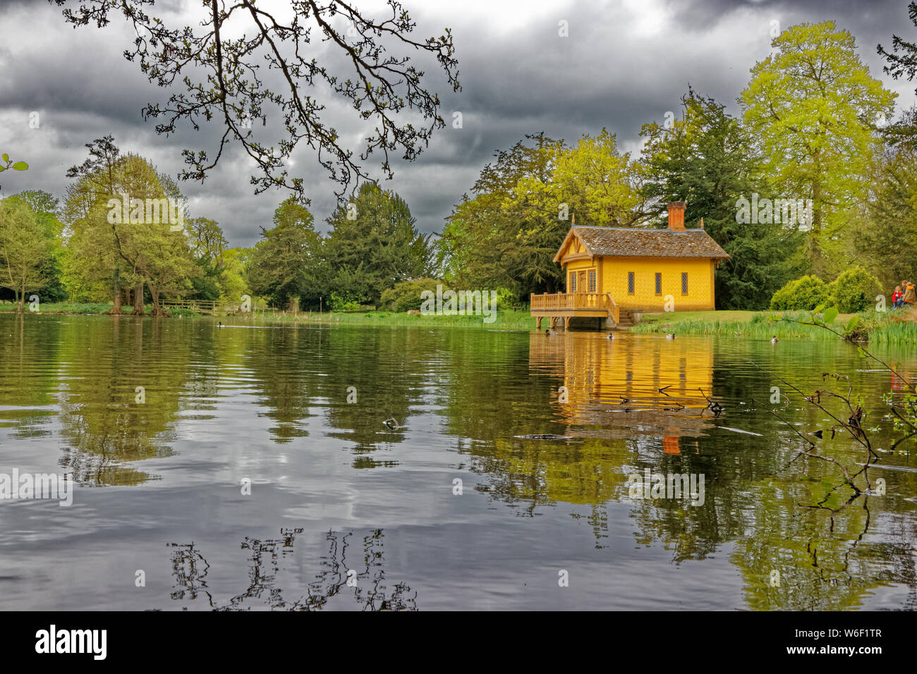 An old recently restored boat house painted yellow overlooking a small lake surrounded by trees with stormy skies above it near Belton in Lincolnshire Stock Photo