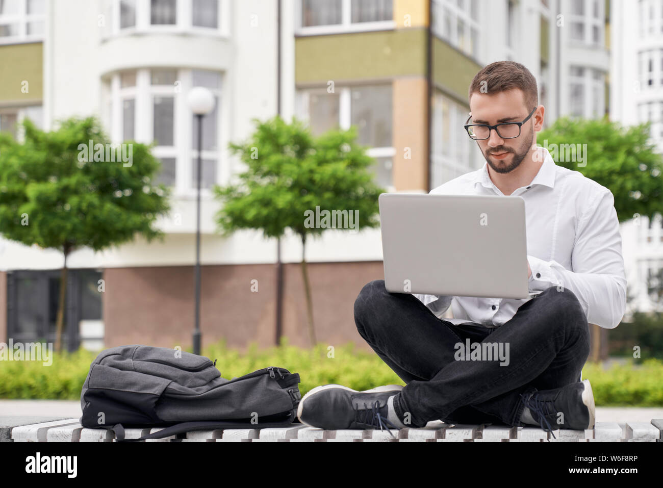Handsome, young bearded man sitting on bench with crossed legs, holding laptop on knees. Stylish student in glasses looking at laptop, working, browsing. Backpack on bench. Stock Photo