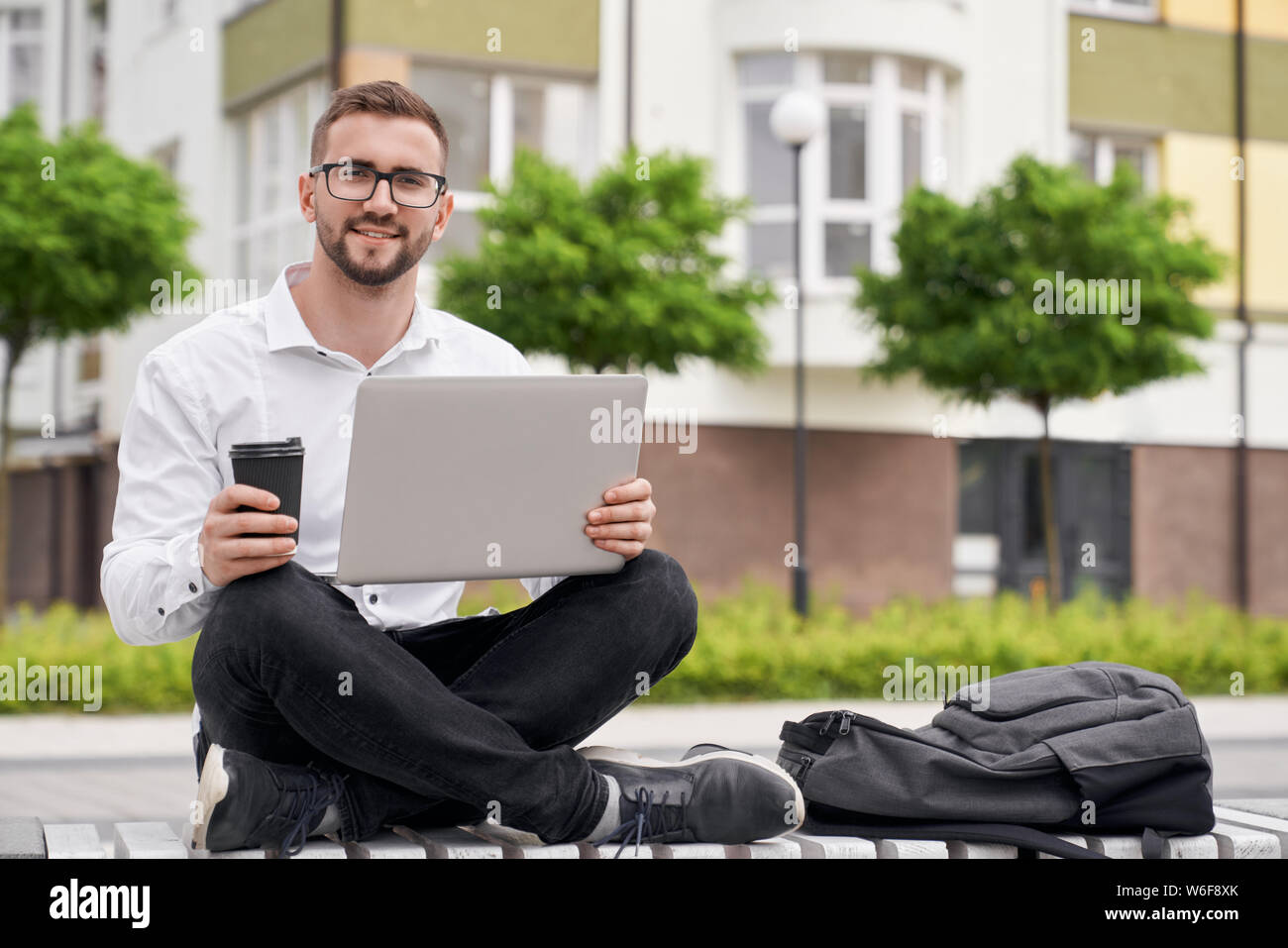 Handsome, positive programmer sitting on bench with crossed legs, holding laptop on knees, looking at camera. Young man in glasses holding coffee cup, posing on background of building. Stock Photo