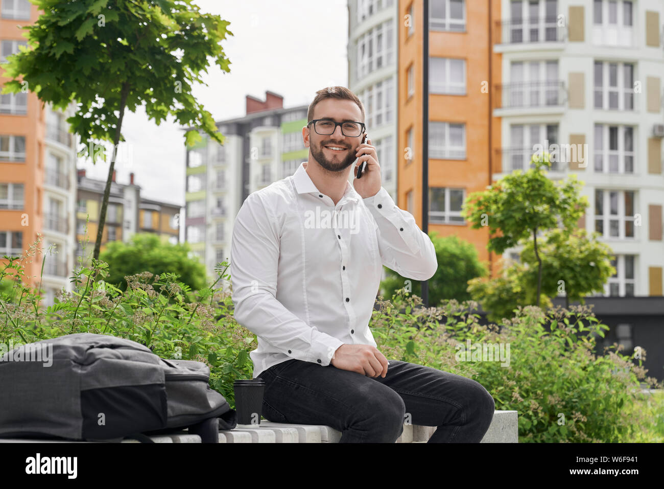 Young freelancer sitting on bench, talking by phone, looking at camera. Handsome man in glasses wearing in white stylish shirt. It worker smiling, posing on background of modern city. Stock Photo