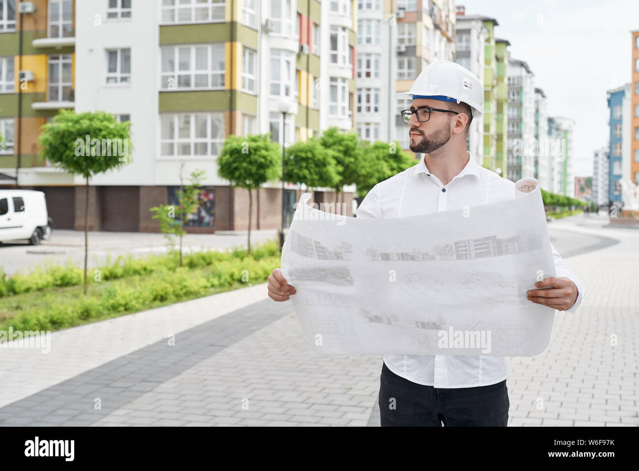 Engineer in white shirt and hardhat holding architectural project on paper, looking at house. Young man in glasses standing outdoors in sleeping area of modern residential development. Stock Photo