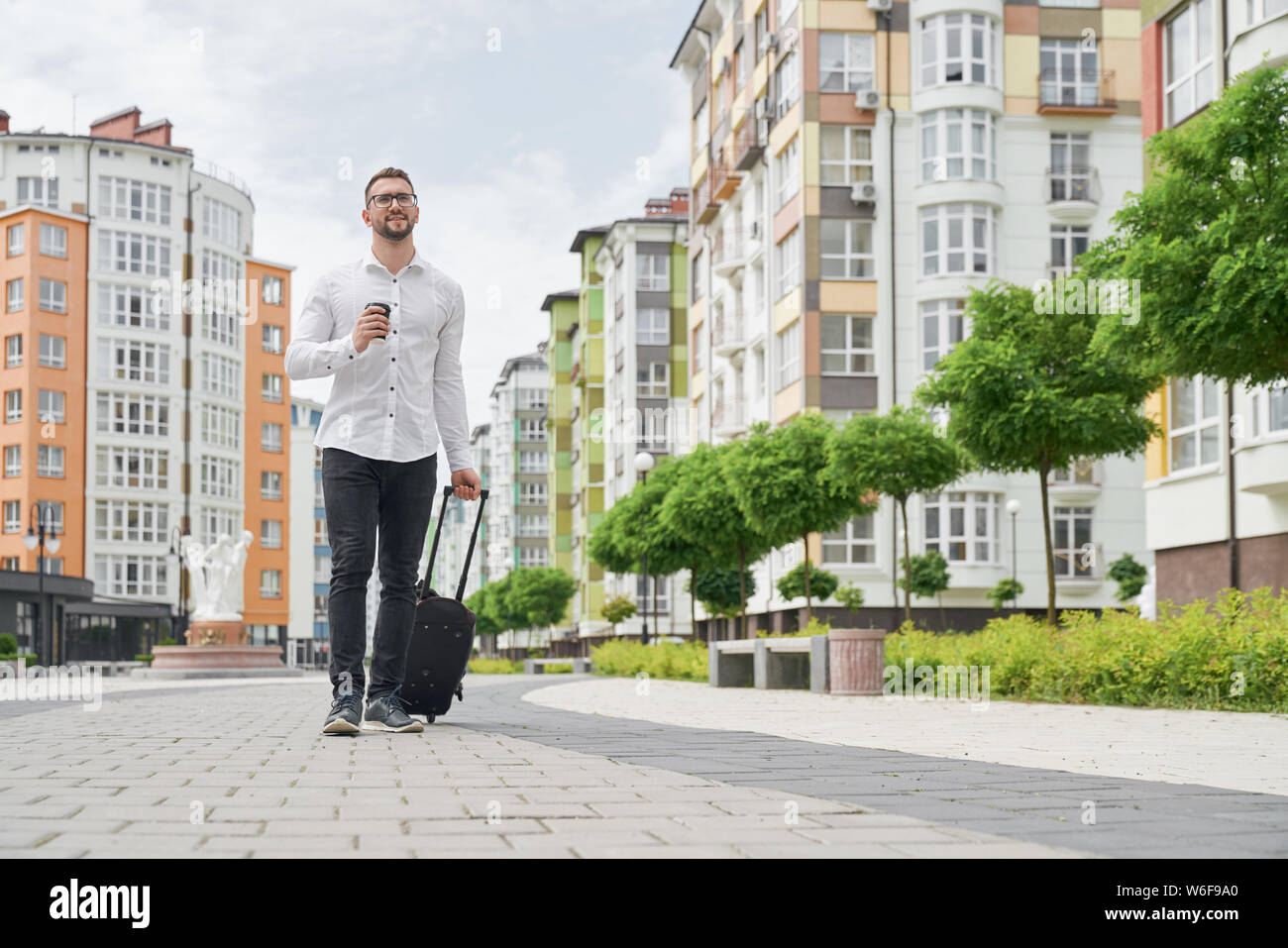 Handsome bearded student in glasses with coffee cup walking down street, looking at building. Young man wearing in white shirt with suitcase on background of multistory houses. Stock Photo