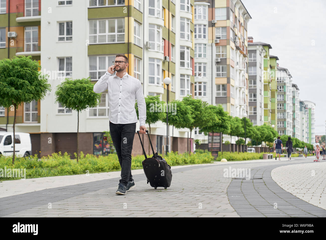 Handsome young man with suitcase walking down street of new apartments buildings. Bearded student in glasses talking by phone, looking at house. Concept of modern city. Stock Photo