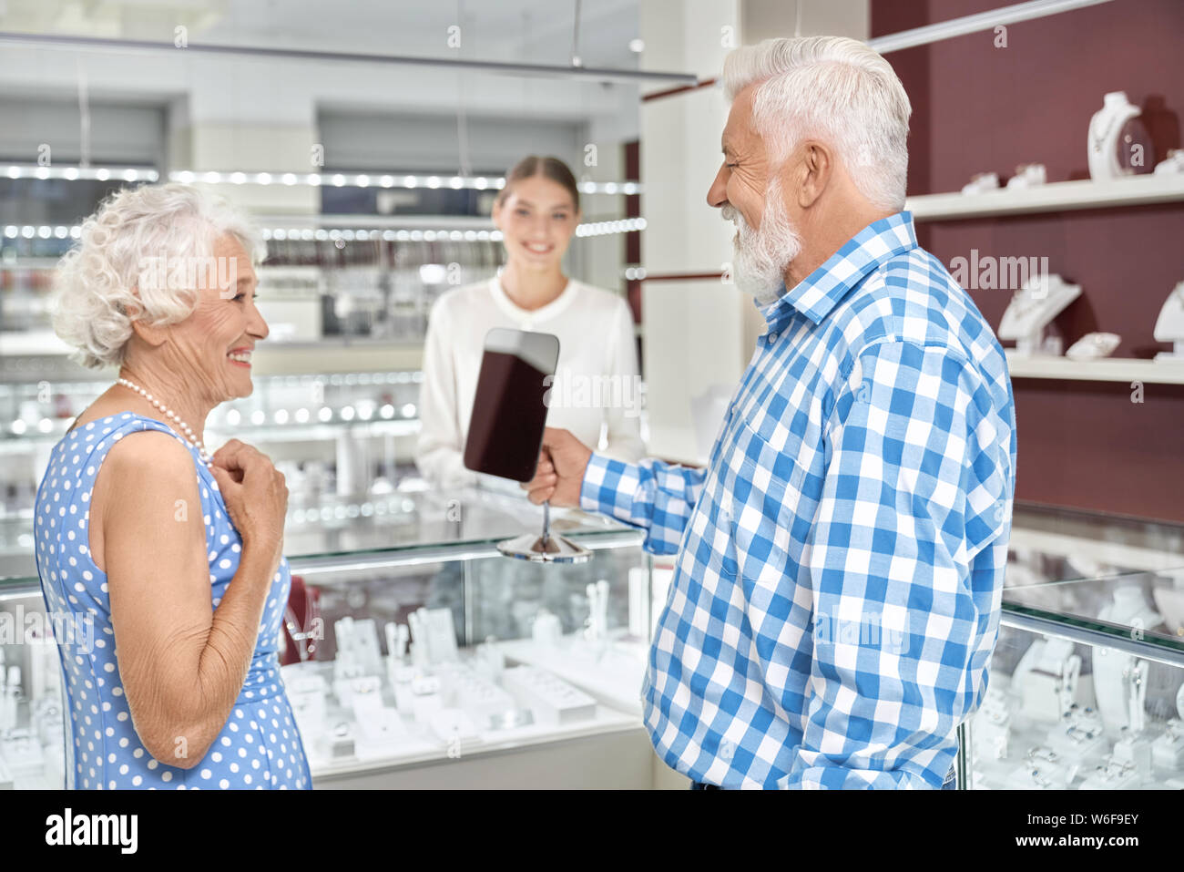 Beautiful elegant aged woman smiling to her husband with grey hair and beard while trying on a pearl necklace at jewelry store. Affectionate man doing luxury present for wife on their anniversary. Stock Photo