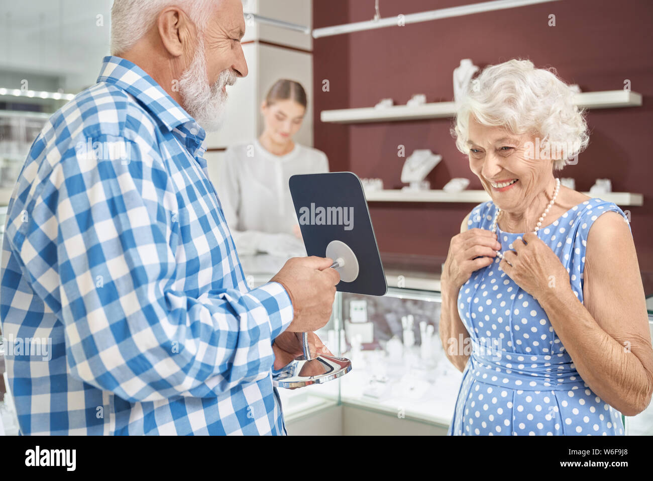 Careful husband with grey hair and beard expressing his love to charming happy wife by buying new luxury necklace at jewelry store. Mature couple enjoying shopping time together Stock Photo