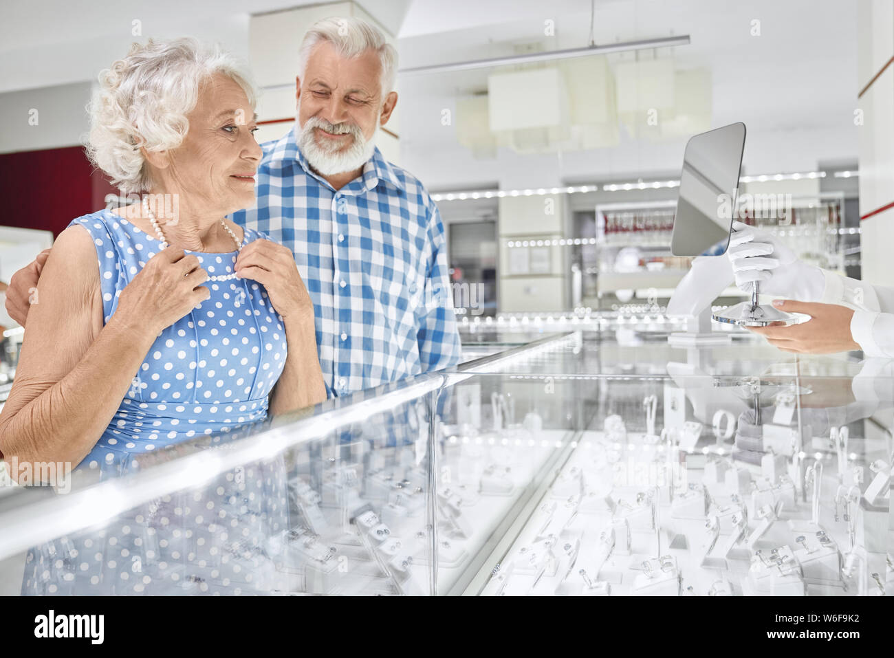 Cheerful bearded senior man in checkered shirt embracing his beautiful wife in blue dress that trying on pearl necklace at jewelry store. Grey haired husband doing pleasant present to beloved woman Stock Photo