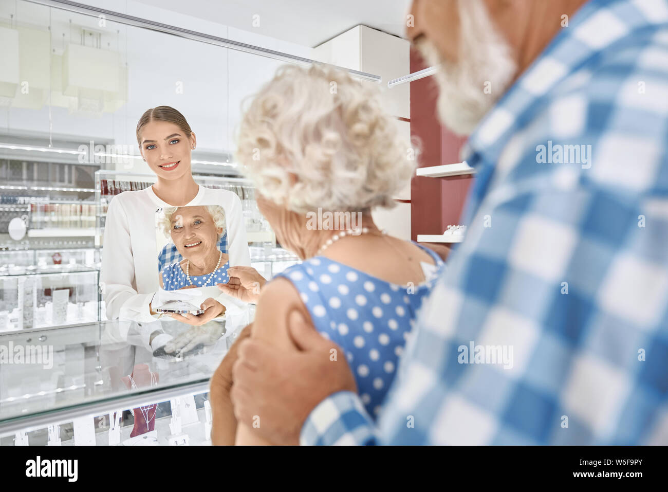 Pretty mature lady in polka dot dress trying on pearl necklace and looking at mirror that keeping attractive smiling female seller. Happy aged wife getting present from her husband at jewelry shop. Stock Photo