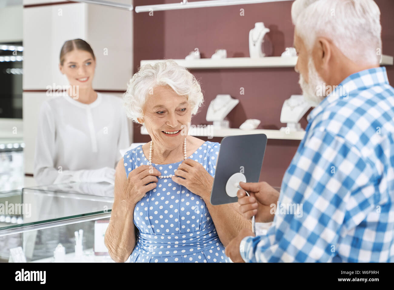 Happy aged woman with grey curly hair in blue trendy dress smiling to her bearded senior husband while choosing luxury pearl necklace at jewelry market. Mature married couple making precious purchase Stock Photo