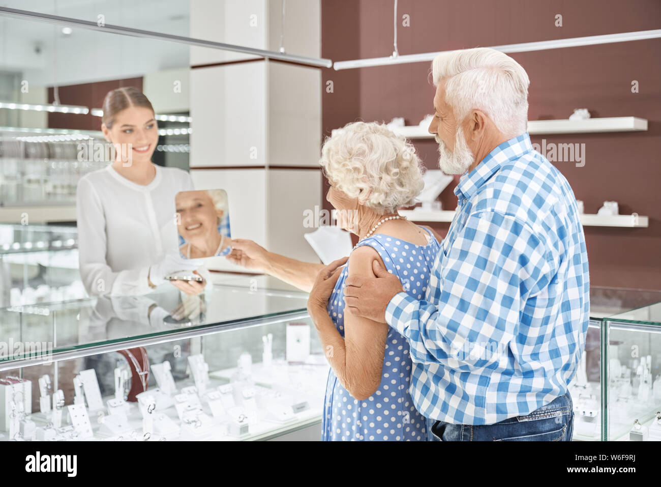 Grey haired man in checkered shirt standing behind his beautiful wife in blue dress embracing her while she trying on amazing pearl necklace at shopping mall. Concept of shopping, sale and consumerism Stock Photo