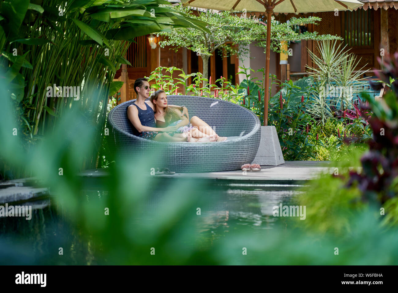 Candid shot of young caucasian couple lounging in modern chair near luxurious pool of tropical hotel and spa in Bali Stock Photo