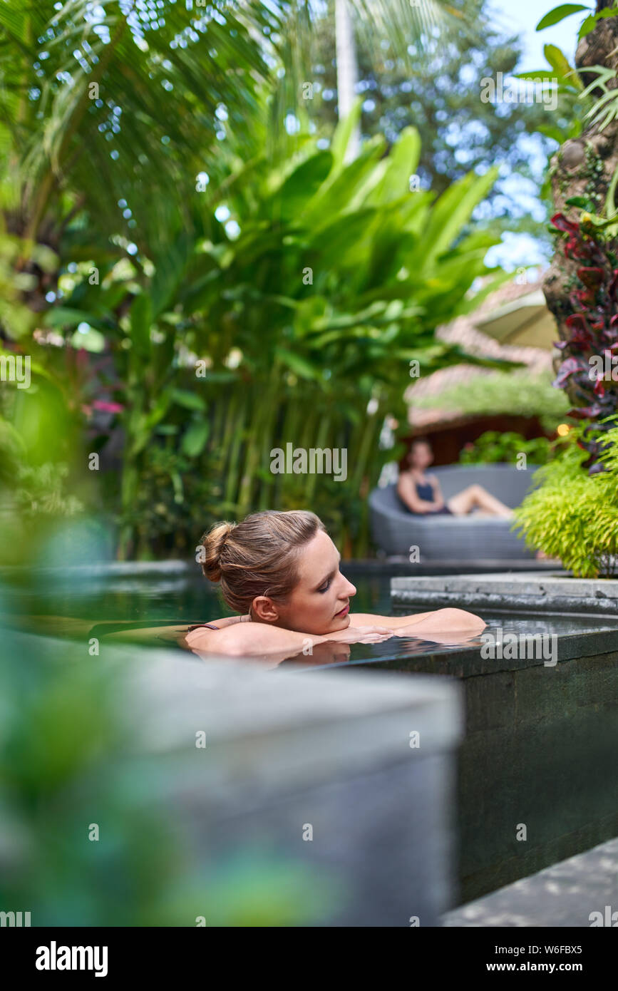 Pretty peaceful woman relaxing outdoors in sunny and lush stone swimming pool of luxurious hotel during tropical holiday in Bali Stock Photo