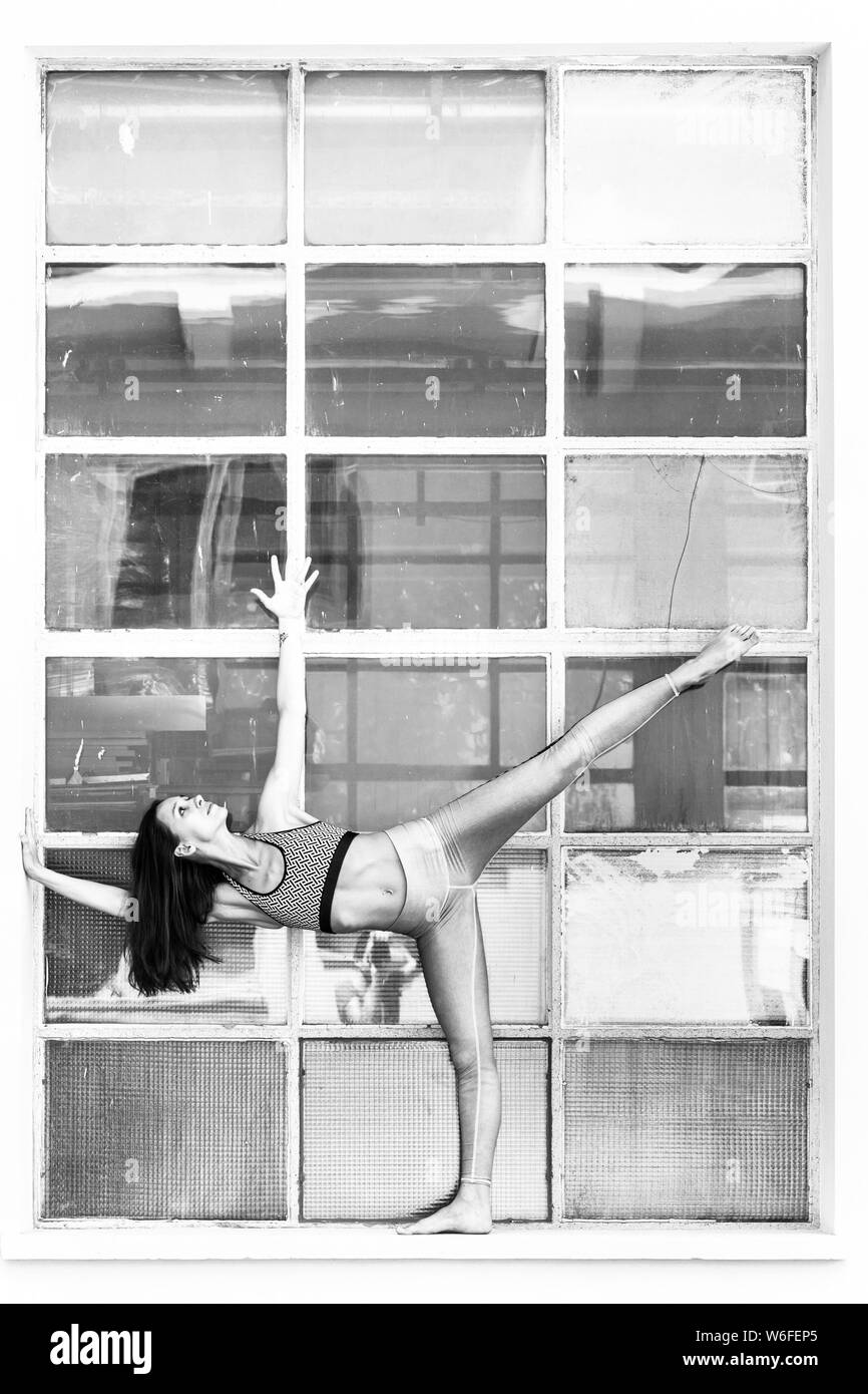 Fit sporty active girl in fashion sportswear doing yoga fitness exercise in front of big industrial window frame. colorful reflections in window glass Stock Photo