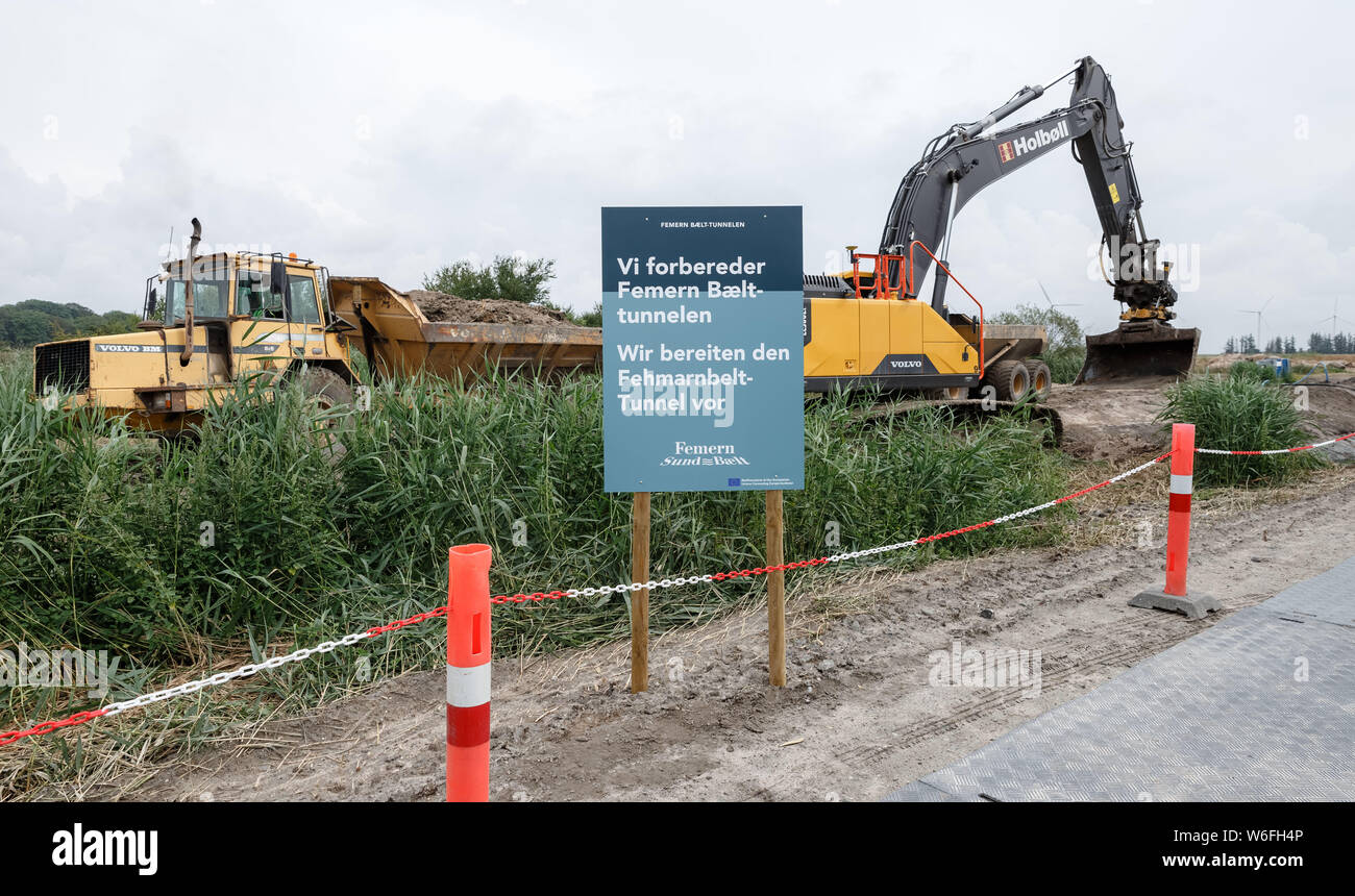 01 August 2019, Denmark, Rødbyhavn: Construction machines dig a drainage ditch behind a sign with the German, Danish inscription 'Wir bereiten den Fehmarnbeltunnel vor' (We are preparing the Fehmarnbelt tunnel) on the future construction site. Photo: Markus Scholz/dpa Stock Photo