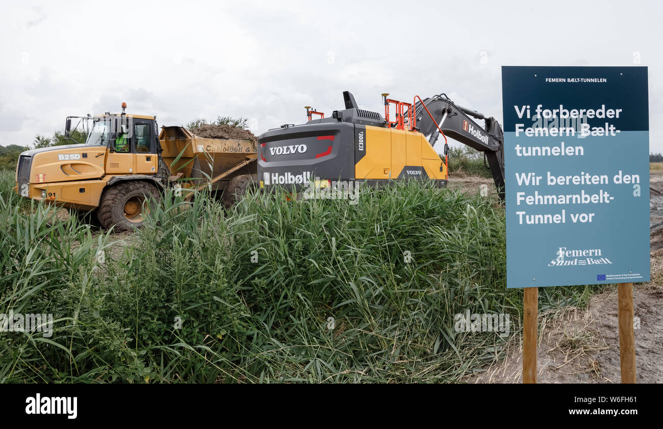 01 August 2019, Denmark, Rødbyhavn: Construction machines dig a drainage ditch behind a sign with the German, Danish inscription 'Wir bereiten den Fehmarnbeltunnel vor' (We are preparing the Fehmarnbelt tunnel) on the future construction site. Photo: Markus Scholz/dpa Stock Photo