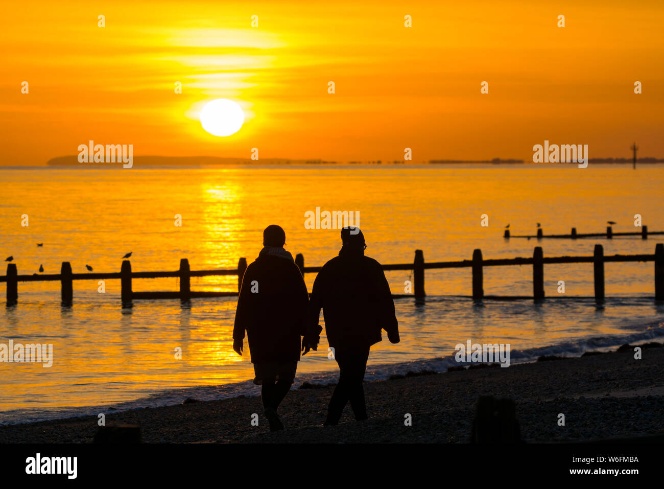 A couple having a romantic walk holding hands along the beach at sunset in Autumn in the UK. Stock Photo