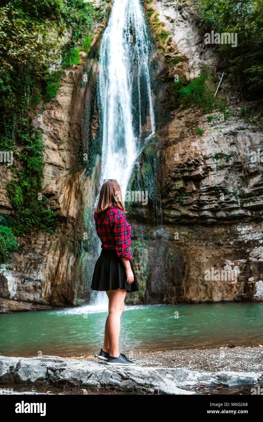 Young brunette woman in red squared shirt and skirt standing near waterfall in park Stock Photo