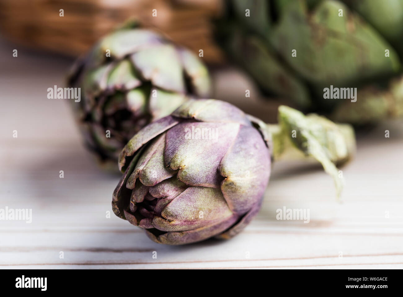 Baby violet artichokes, whole on wooden table with basket in background. Stock Photo