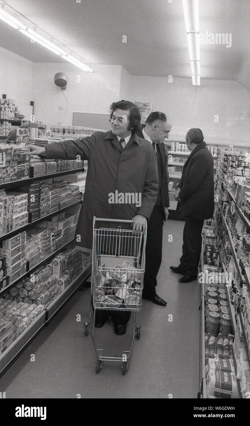 1970s, historical, a man in a raincoat using a metal trolley to do his shopping in a supermarket, England, UK. Stock Photo