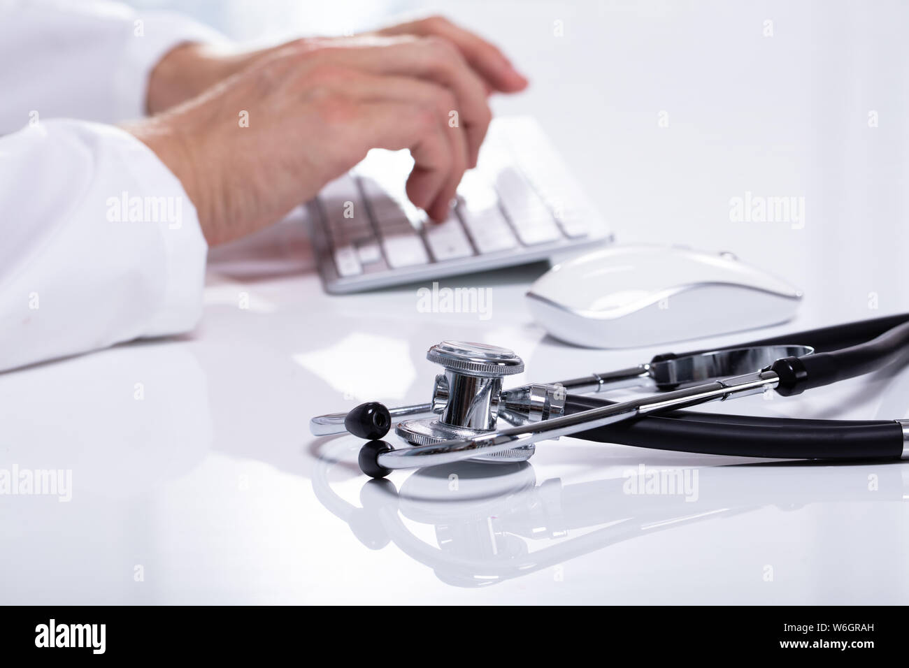 Close-up Of Stethoscope On White Desk Besides Doctor Typing On Wireless Keyboard Stock Photo