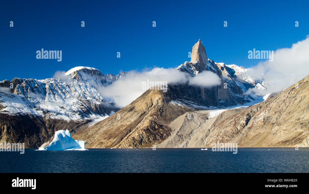 Exploring Greenland’s fjord systems, I stumbled upon this wild and immense landscape which immediately stole my heart. The singular pinnacle of rock nestled among blue skies, was framed by blankets of cloud, and dwarfed passing icebergs which paraded past. It was like nothing I’d ever seen before.  Notes: From the deck of a ship Stock Photo