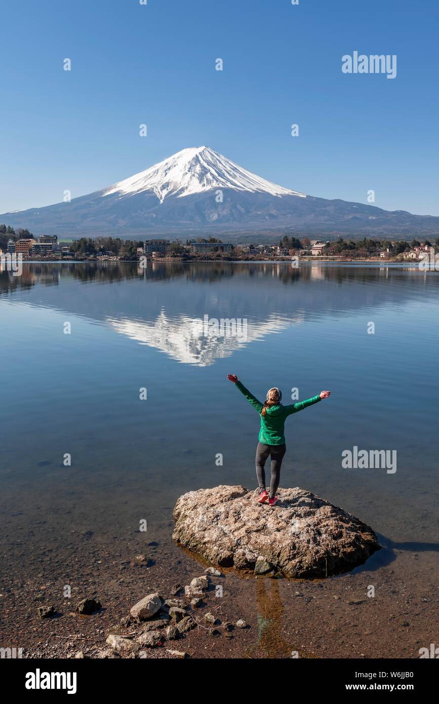 Young woman stands on stone in the water and stretches arms into the air, Lake Kawaguchi, back volcano Mt. Fuji, Yamanashi Prefecture, Japan Stock Photo