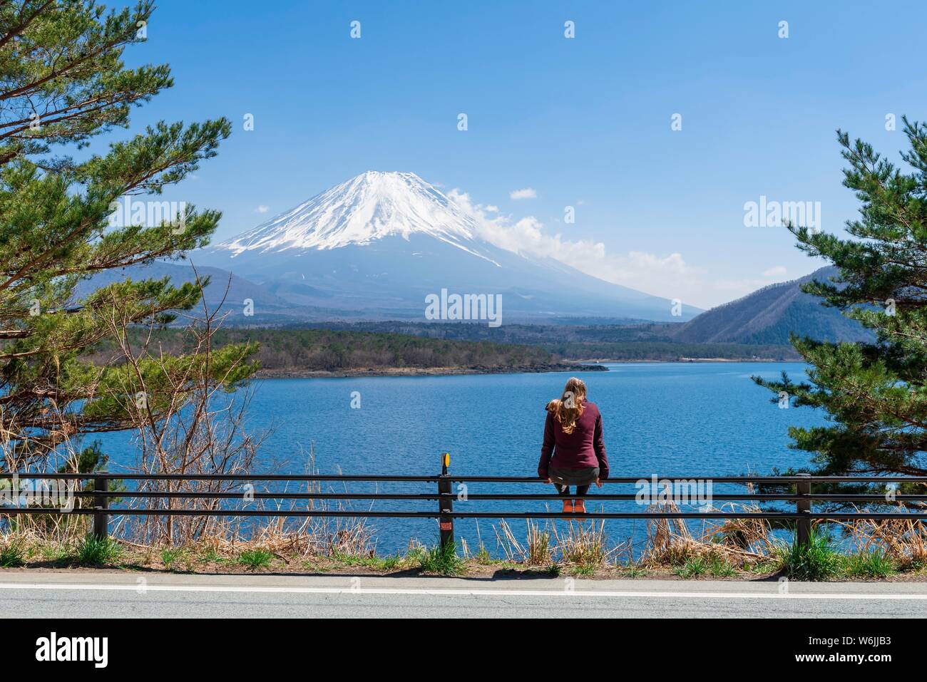 Young woman sitting on a railing next to a road and looking across the lake to the volcano Mt Fuji, Motosu Lake, Yamanashi Prefecture, Japan Stock Photo