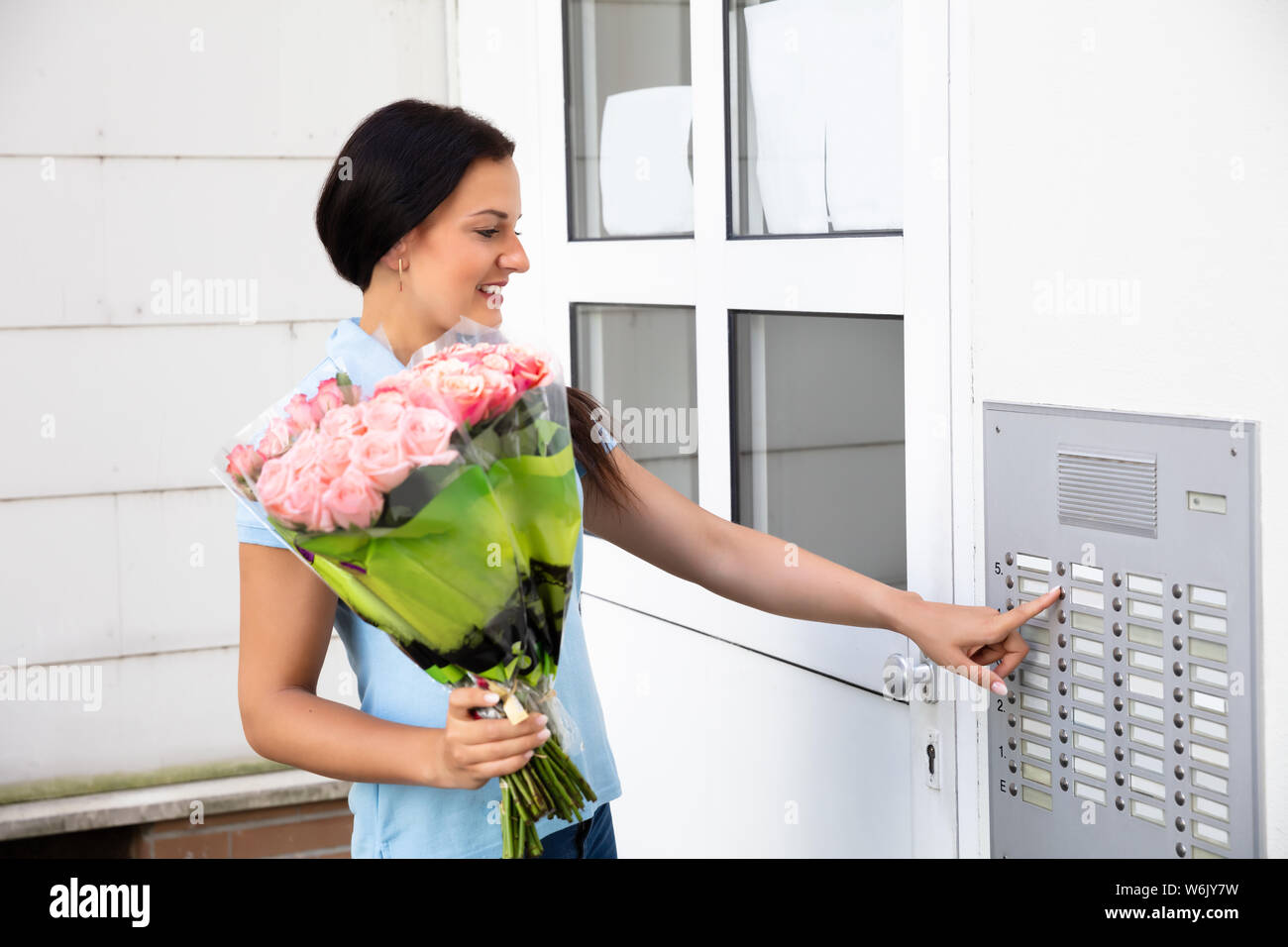 Happy Young Woman Holding Bouquet Pressing Button Of Intercom To Enter Home Stock Photo